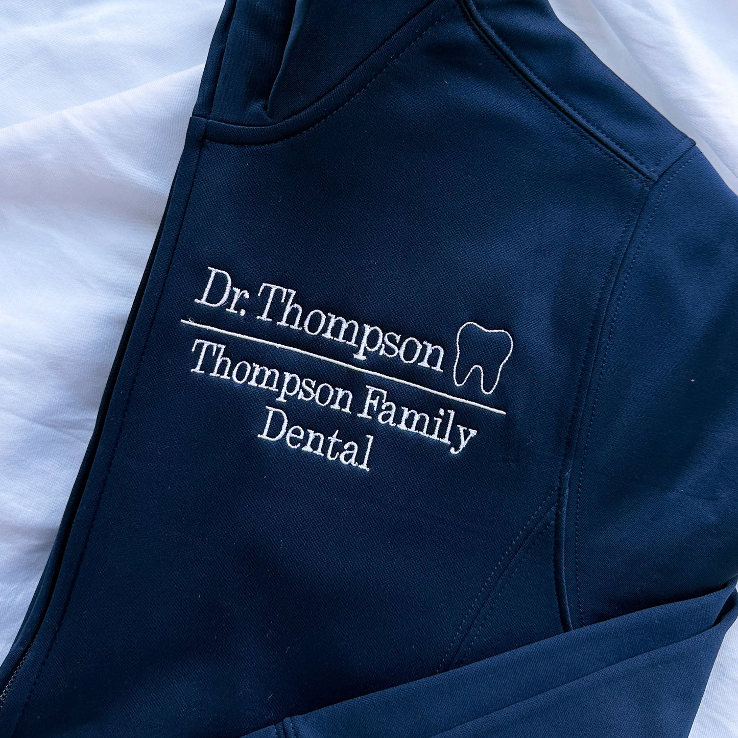 close up image of embroidery on a navy full zip polyester jacked that has Dr. Thompson with mini outline tooth on the first line and on a line below Thompson Family Dental