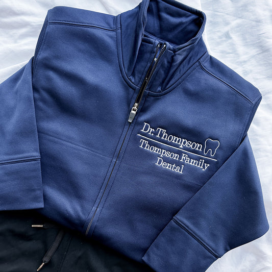 Navy full zip polyester dental jacket with white embroidered design on the left chest. The embroidery showcases Dr. Thompson with mini outline tooth on the first line and on a line below Thompson Family Dental