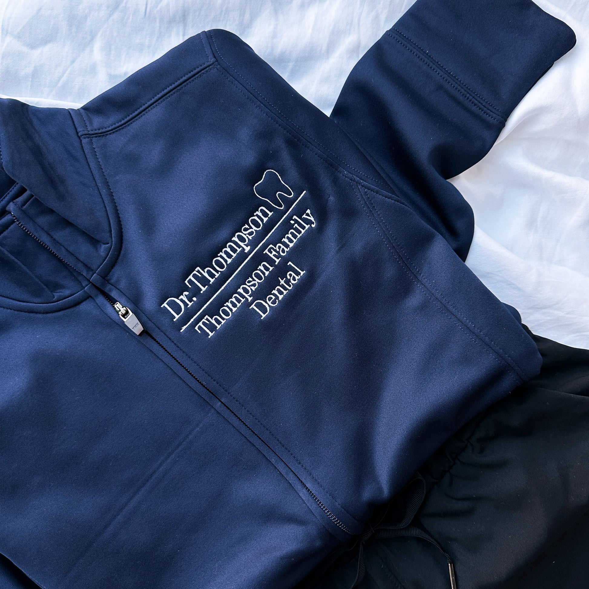 close up image of embroidery on a navy full zip polyester jacked that has Dr. Thompson with mini outline tooth on the first line and on a line below Thompson Family Dental