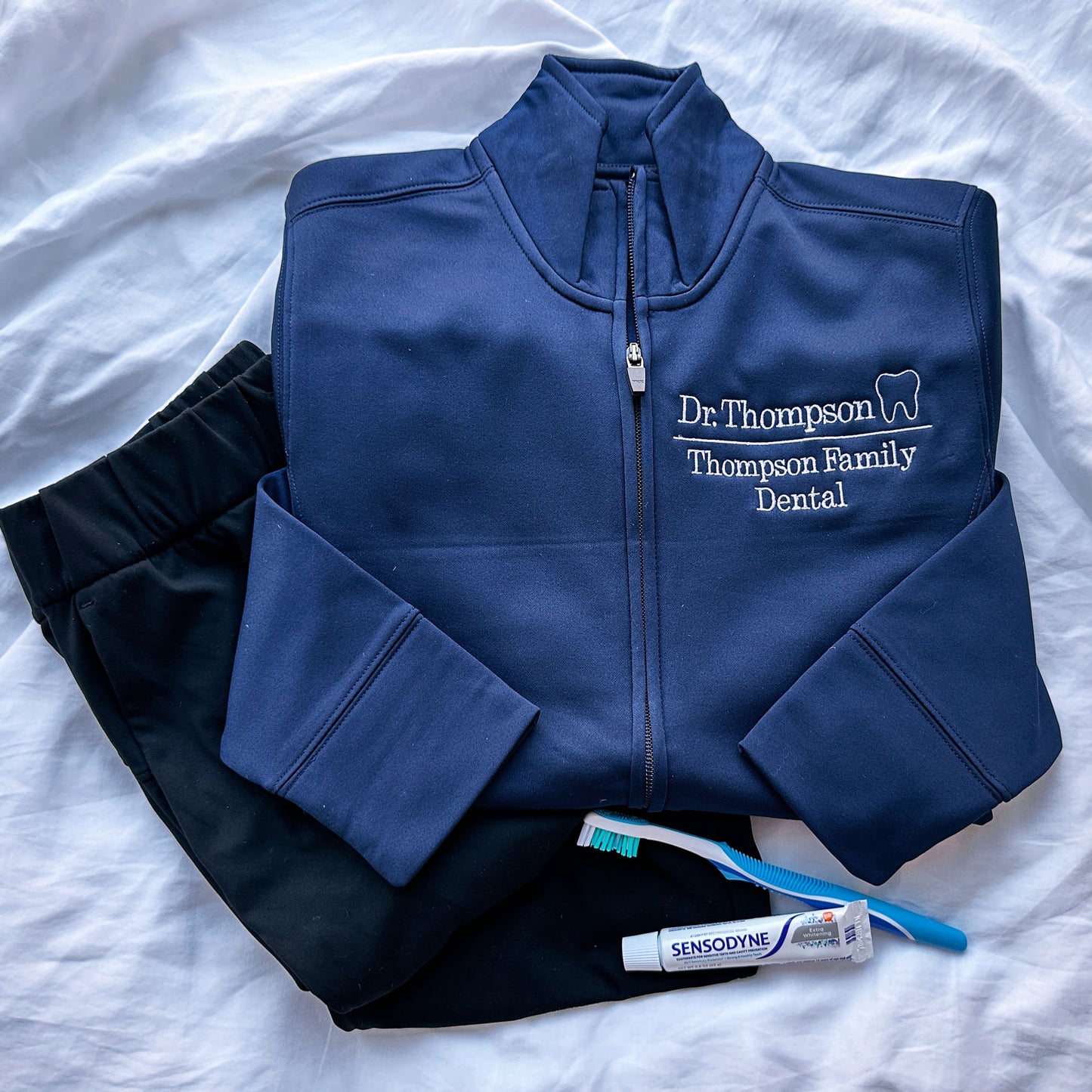 Navy full zip polyester dental jacket with white embroidered design on the left chest. The embroidery showcases Dr. Thompson with mini outline tooth on the first line and on a line below Thompson Family Dental styled with scrubs and a tooth brush and tooth paste