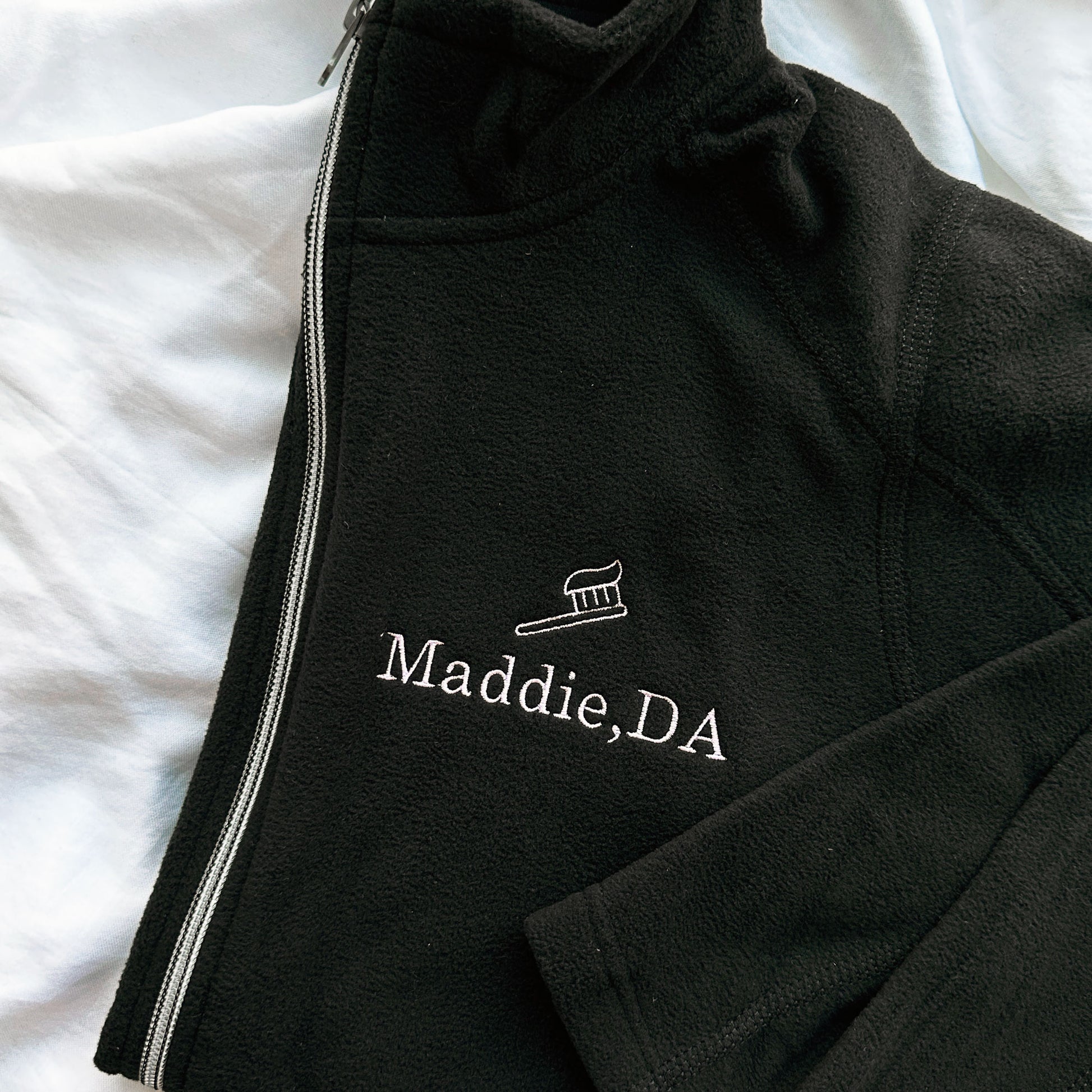 close up of a full zip fleece black jacket with mini embroidered toothbrush outline and name Maddie DA in lilac thread on the left chest