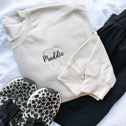 Sand crewneck sweatshirt styled with sneakers and scrub pants. on the left chest is embroidered a white tooth outline with name maddie in script and black thread over the outlined tooth.
