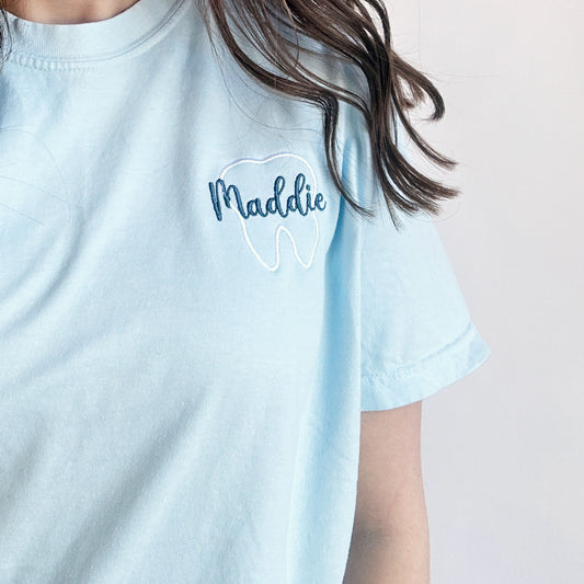 close up of a chambray comfort colors short sleeve tee with embroidered white tooth outline and name Maddie in a script  font and French blue thread over the tooth