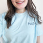 young woman wearing  a chambray comfort colors short sleeve tee with embroidered white tooth outline and name Maddie in a script font and French blue thread over the tooth on the left chest