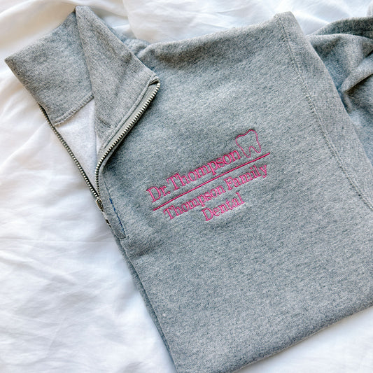 flat lay of an oxford quarter zip sweatshirt. On the left chest in embroidered Dr. Thompson Thompson Family Dental and mini outline tooth in pink thread