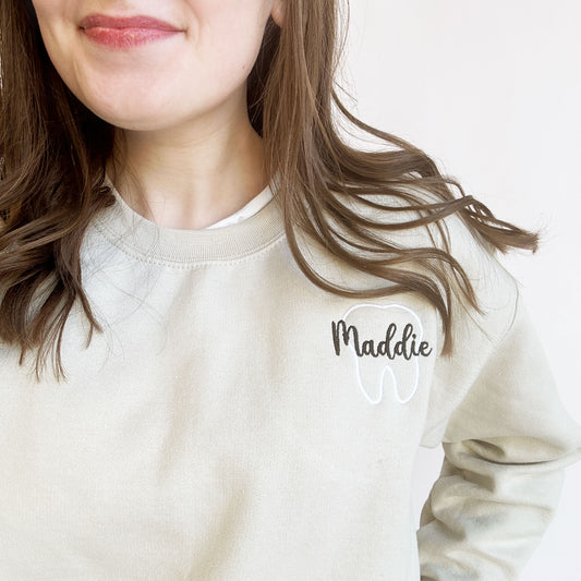 close up of a young woman wearing a sand crewneck sweatshirt white tooth outline with name maddie in script and black thread over the outlined tooth.