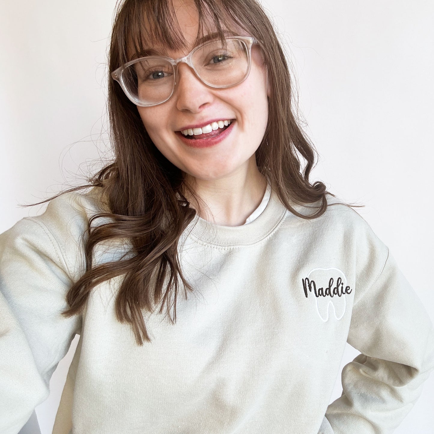 young woman wearing a sand crewneck sweatshirt white tooth outline with name maddie in script and black thread over the outlined tooth.