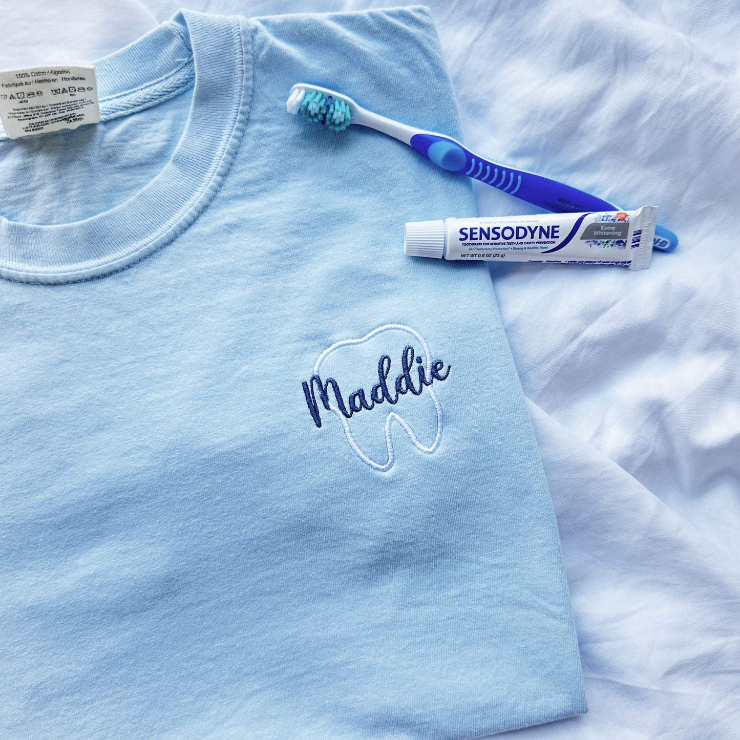 close up of a chambray comfort colors short sleeve tee with embroidered white tooth outline and name Maddie in a script  font and French blue thread over the tooth styled with a tooth brush and tooth paste