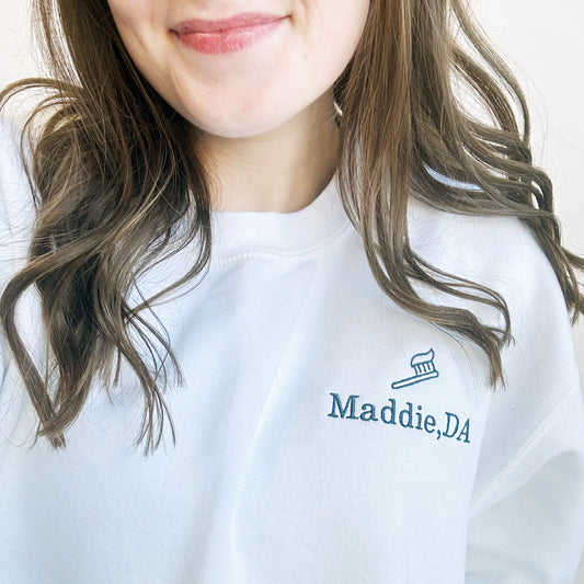 Close up of young woman wearing a white crewneck sweatshirt with maddie DA and l=mini tooth brush design embroidered on the left chest in white thread