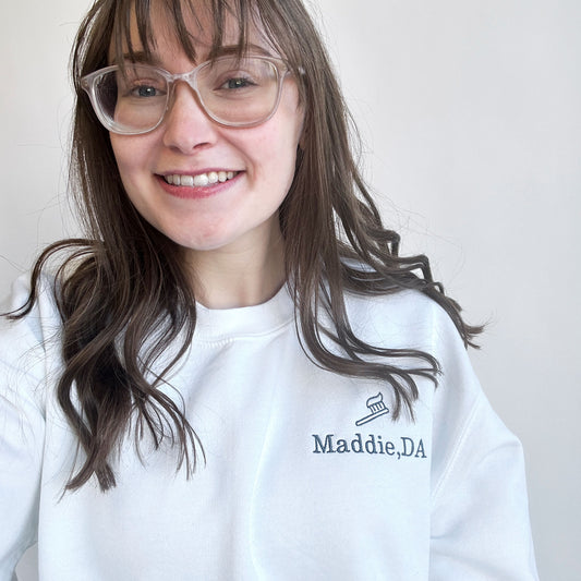 Young woman wearing a white crewneck sweatshirt with maddie DA and l=mini tooth brush design embroidered on the left chest in white thread