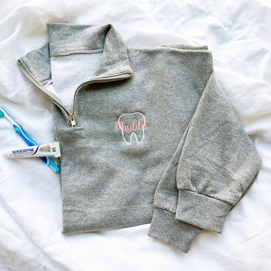 up close of an oxford quarterzip sweatshirt with white outline tooth embroidery and name Maddie in script font and coral pink thread embroidered over the tooth