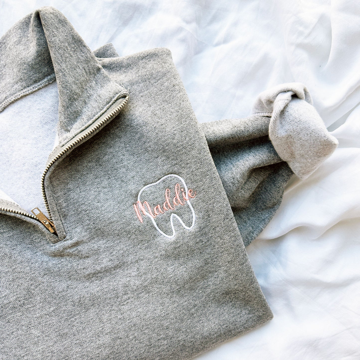 up close of an oxford quarterzip sweatshirt with white outline tooth embroidery and name Maddie in script font and coral pink thread embroidered over the tooth