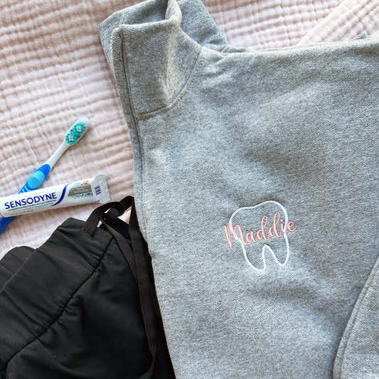 oxford quarterzip sweatshirt with white outline tooth embroidery and name Maddie in script font and coral pink thread embroidered over the tooth styled with black scrubs, a tooth brush and tooth paste