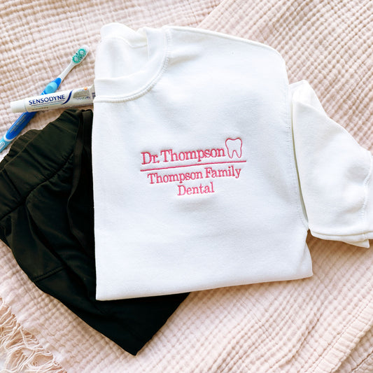 flat lay of white crewneck sweatshirt with embroidered dr. thompson, thompson family dental in pink with mini outline tooth on the left chest styled with scrubs and a tooth brush and tooth paste