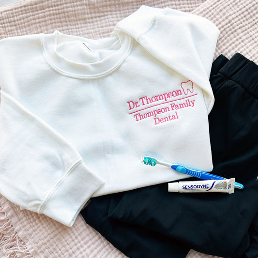 flat lay of white crewneck sweatshirt with embroidered dr. thompson, thompson family dental in pink with mini outline tooth on the left chest