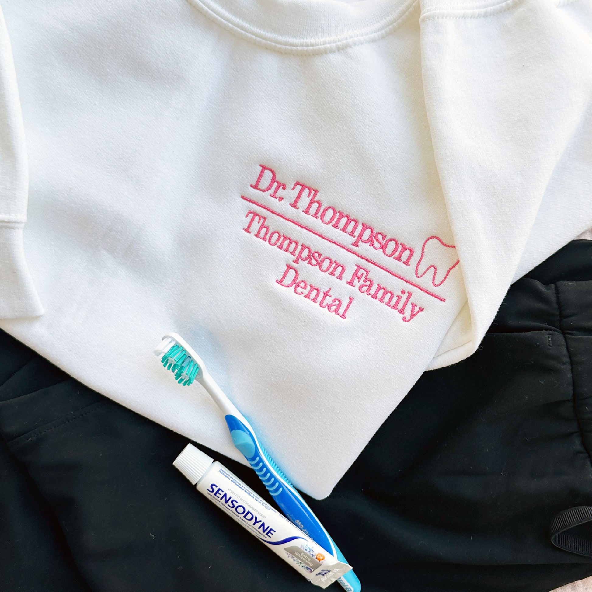 close up of a flat lay of white crewneck sweatshirt with embroidered dr. thompson, thompson family dental in pink with mini outline tooth on the left chest