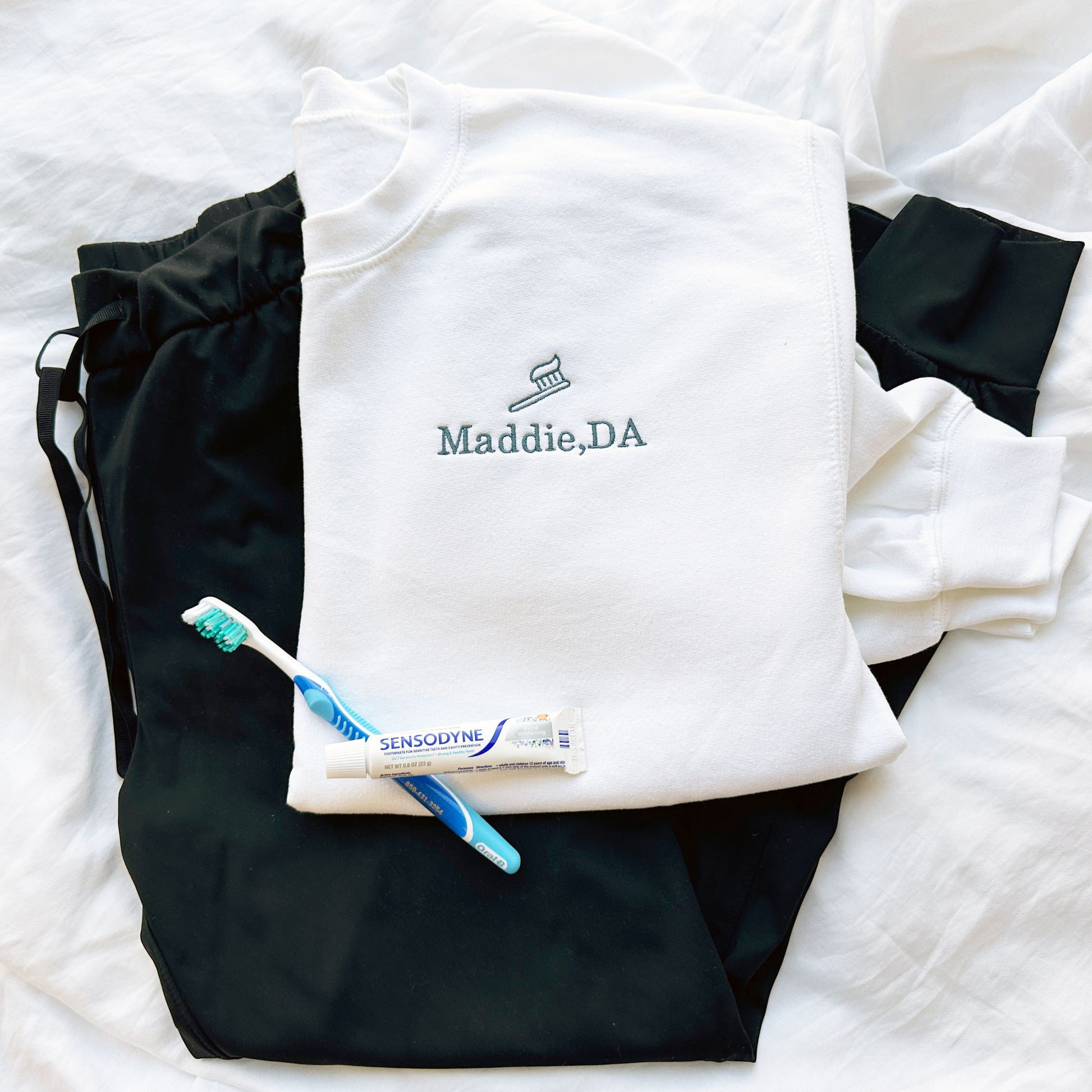 flat lay of a white crewneck sweatshirt, lack scrubs, and toothbrush and tooth paste. Embroidered on the left chest is a toothbrush/toothpaste outlined design above the Name Maddie, DA in French blue threads