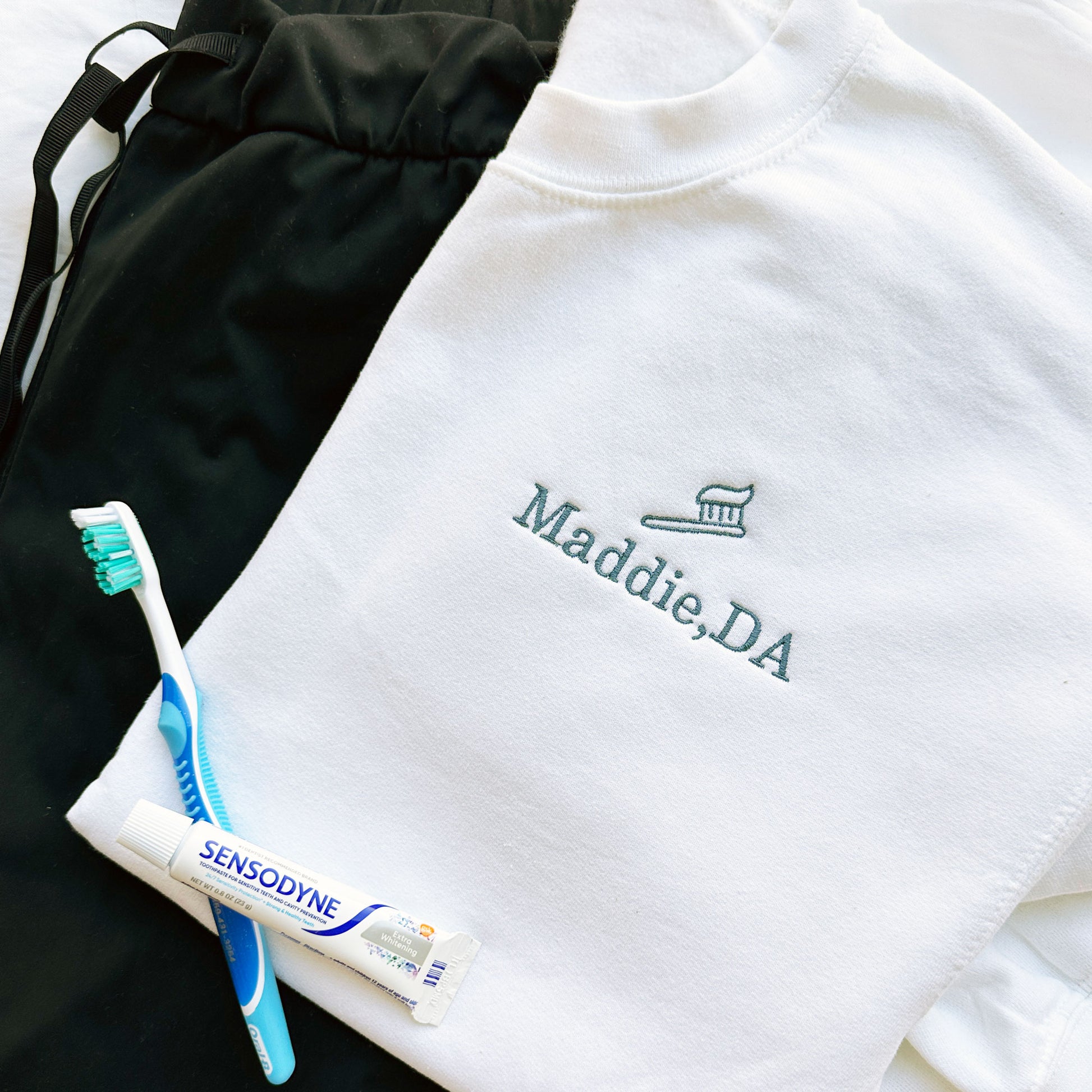 close up of a flat lay of scrubs and tooth brush and tooth paste with white crewneck sweatshirt and mini outline toothbrush embroidered design with name Maddie, DA underneath embroidered in French blue thread