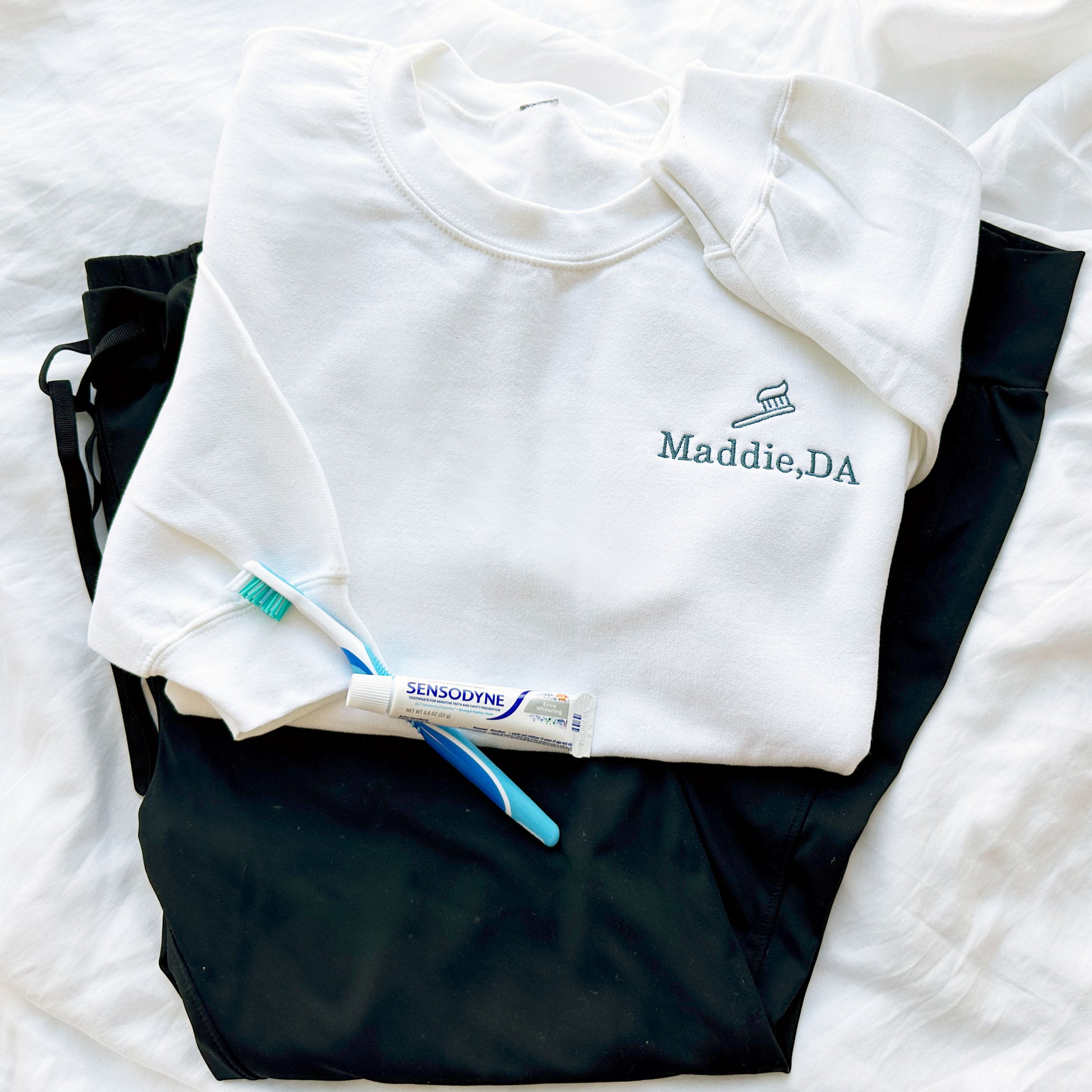flat lay of a white crewneck sweatshirt, lack scrubs, and toothbrush and tooth paste. Embroidered on the left chest is a toothbrush/toothpaste outlined design above the Name Maddie, DA in French blue threads