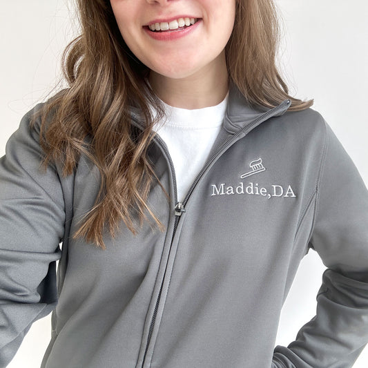 young woman wearing a gray polyester full zip jacket with custom name and toothbrush embroidered design on the left chest
