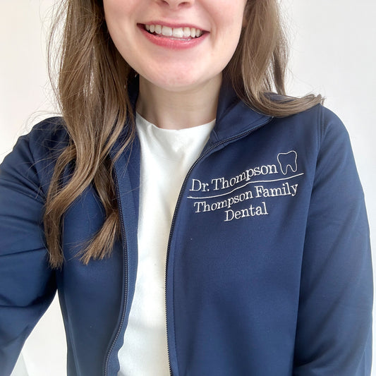 young woman wearing a Navy full zip polyester dental jacket with white embroidered design on the left chest. The embroidery showcases Dr. Thompson with mini outline tooth on the first line and on a line below Thompson Family Dental