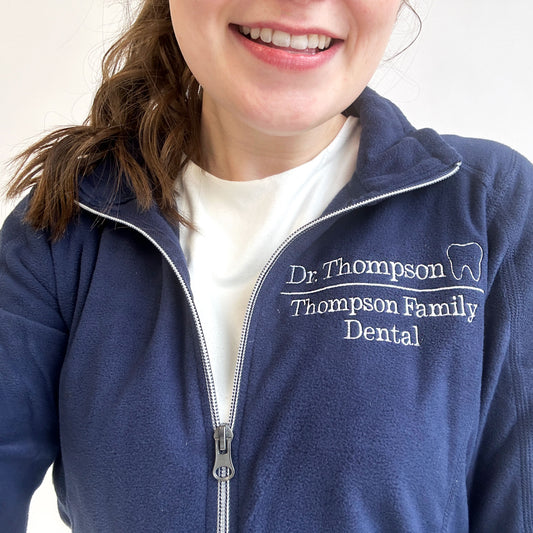 young woman wearing a navy fleece jacket with Dr, thompson Thompson family dental and mini outline tooth embroidered on the left chest on white thread