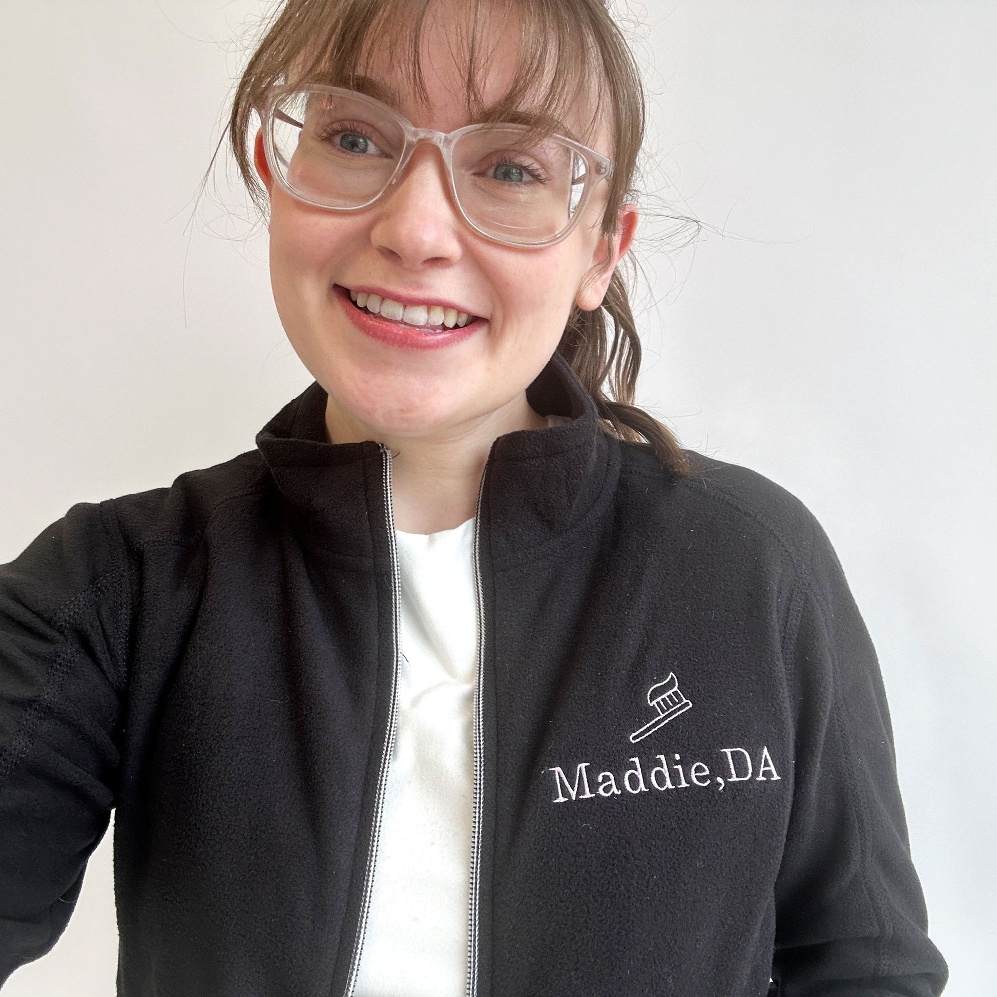 young woman wearing a full zip fleece black jacket with mini embroidered toothbrush outline and name Maddie DA in lilac thread on the left chest