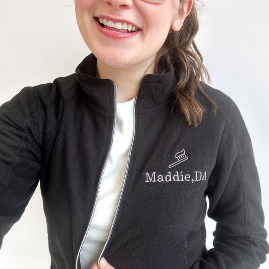 young woman wearing a full zip fleece black jacket with mini embroidered toothbrush outline and name Maddie DA in lilac thread on the left chest