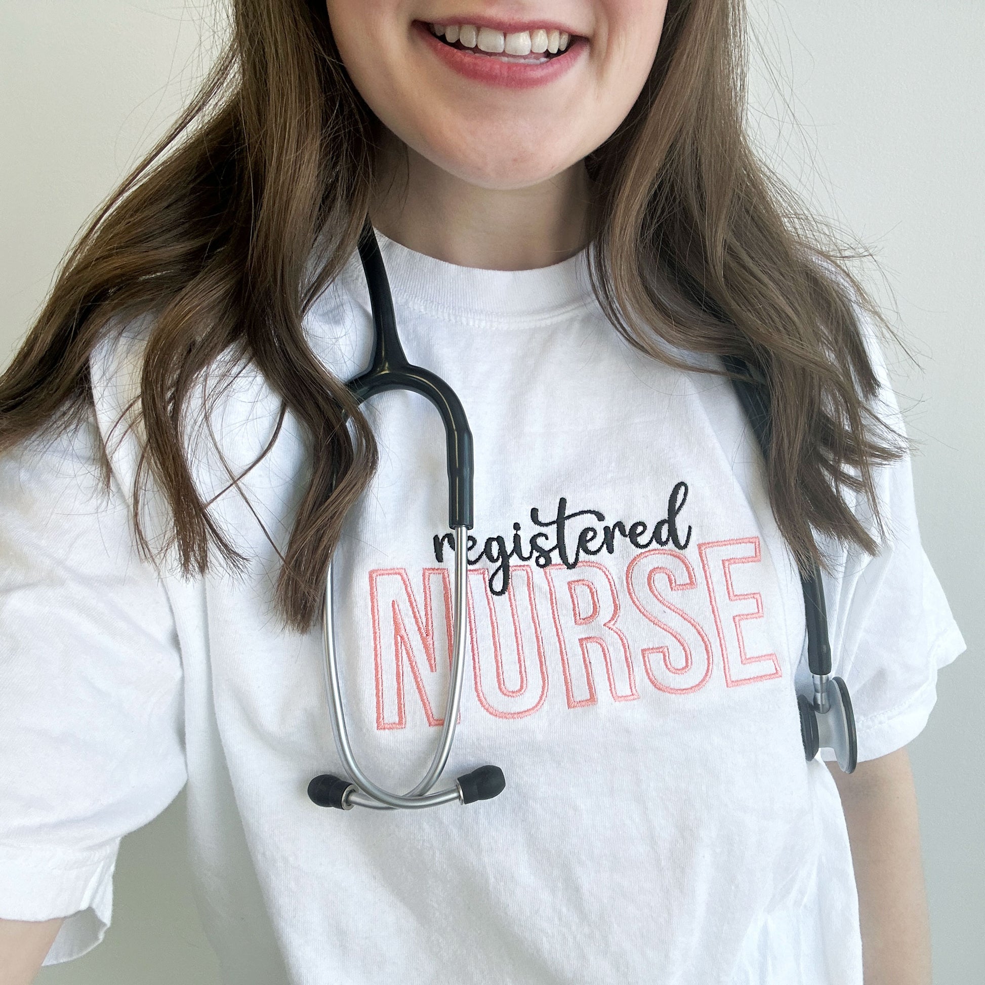 close up of a Young woman wearing a white comfort colors t-shirt with custom text registered nurse embroidered design across the chest in black and coral pink threads.
