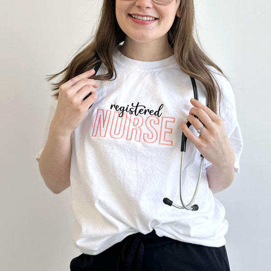 Young woman wearing a white comfort colors t-shirt with custom text registered nurse embroidered design across the chest in black and coral pink threads. 