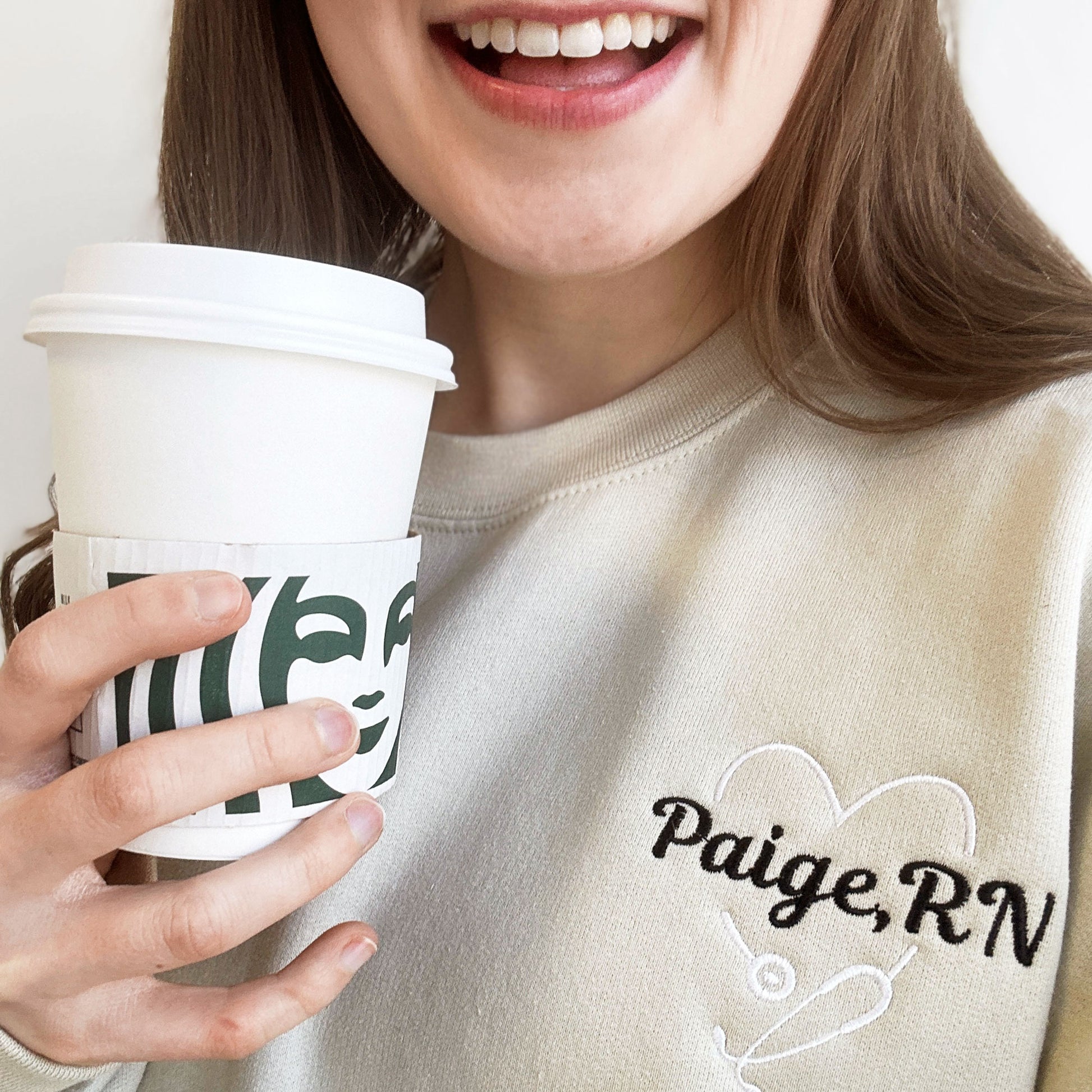 woman wearing a sand crewneck sweatshirt with a custom large heart embroidered stethoscope with her name embroidered through the center in a script font