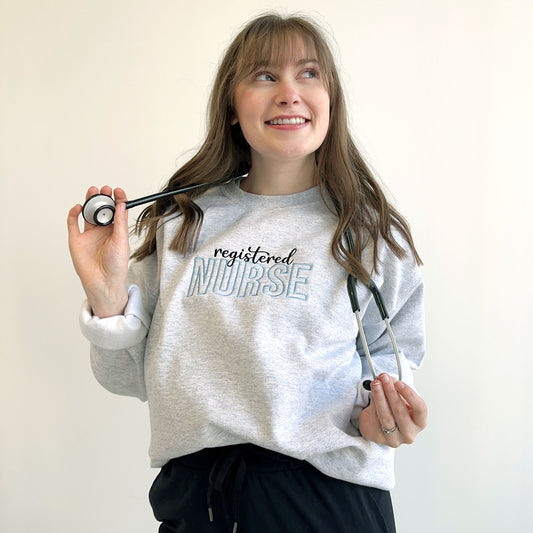 young woman modeling an ash crewneck sweatshirt with custom embroidered specialty and nurse design across the chest on baby blue an black threads