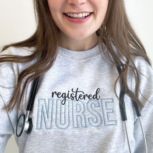 close up of a young woman wearing an ash crewneck sweatshirt with registered nurse embroidered design in black and baby blue threads