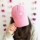 Woman wearing a pink baseball hat with a mini white embroidered heart 