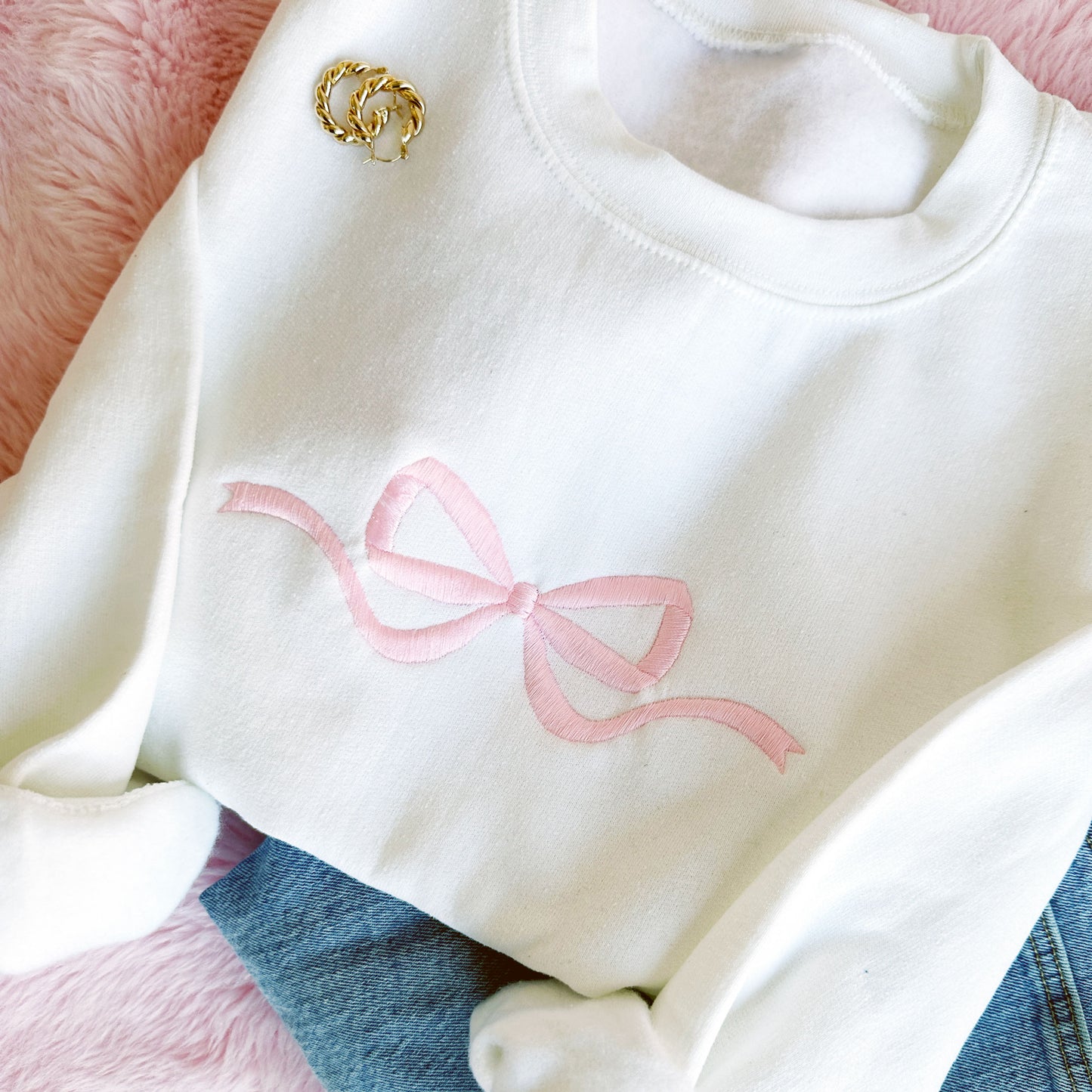 styled flat lay of embroidered bow design on a white crewneck sweatshirt with earrings and jeans in the background