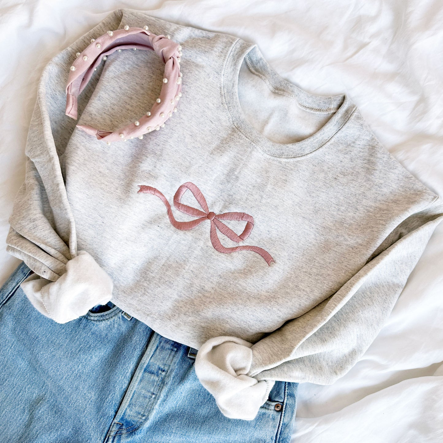 styled flat lay of a heather oatmeal crewneck sweatshirt with mauve embroidered bow