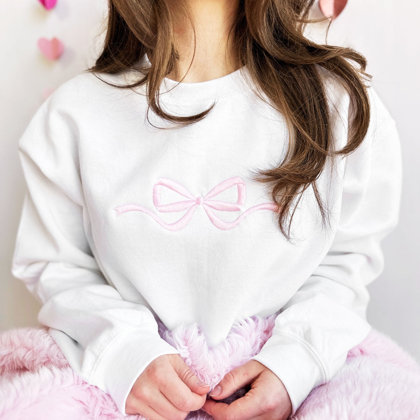 woman wearing a white crewneck sweatshirt with baby pink embroidered bow across the chest