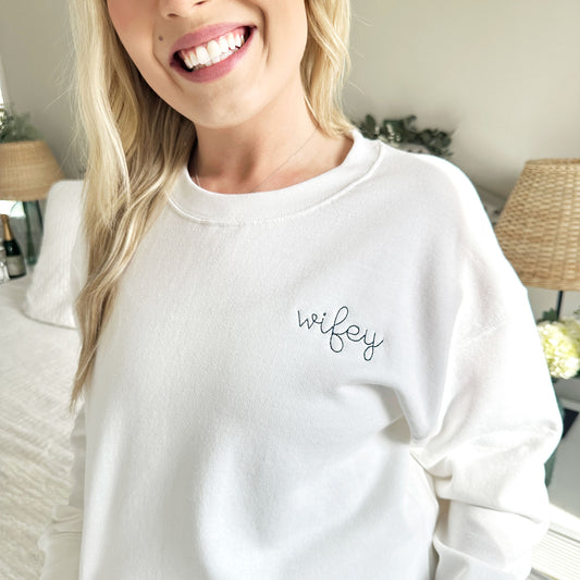 close up of a young woman wearing a white crewneck sweatshirt with wifey embroidered in small script font in French blue thread