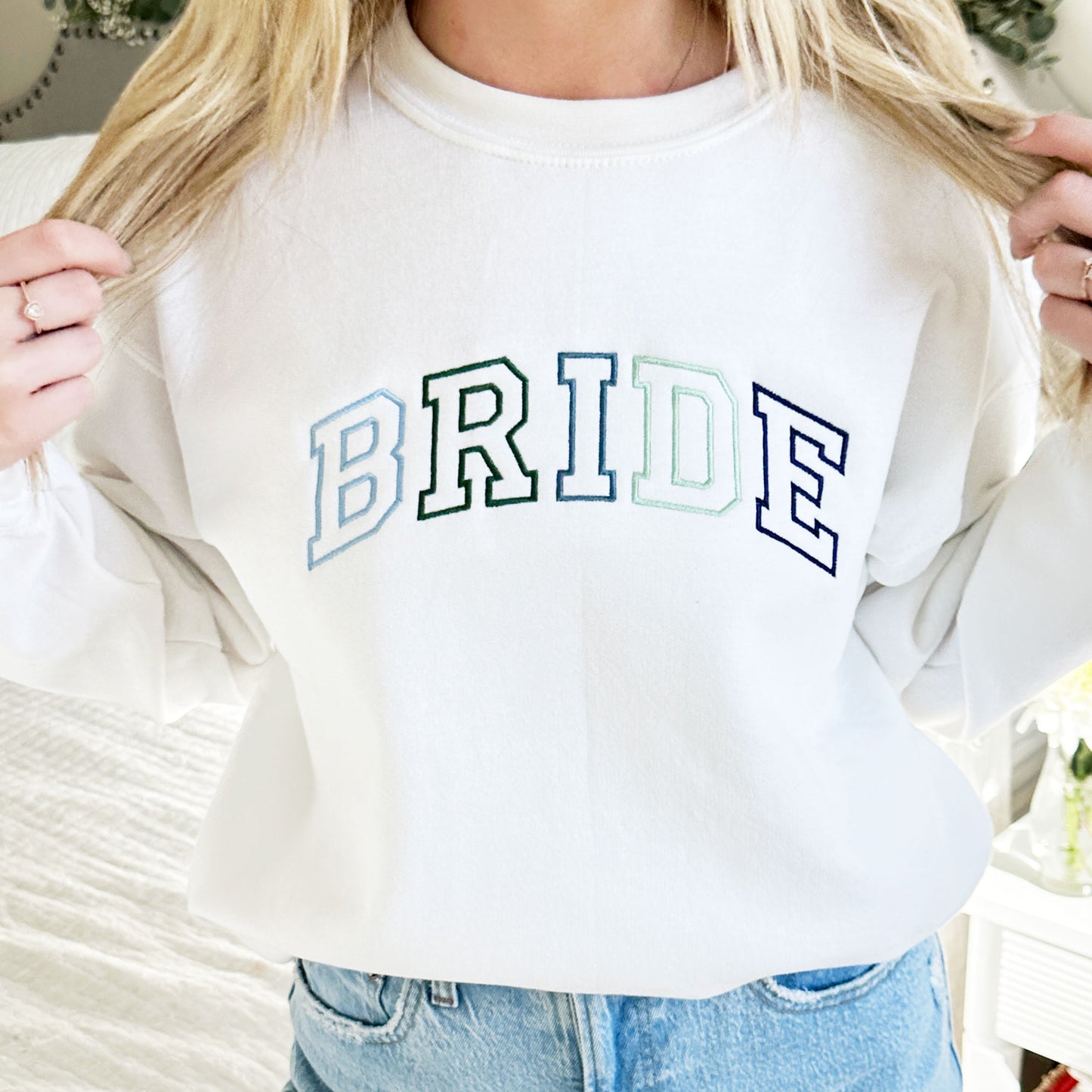 woman wearing a white crewneck sweatshirt with a custom large bride design embroidered in blue and green thread across the chest