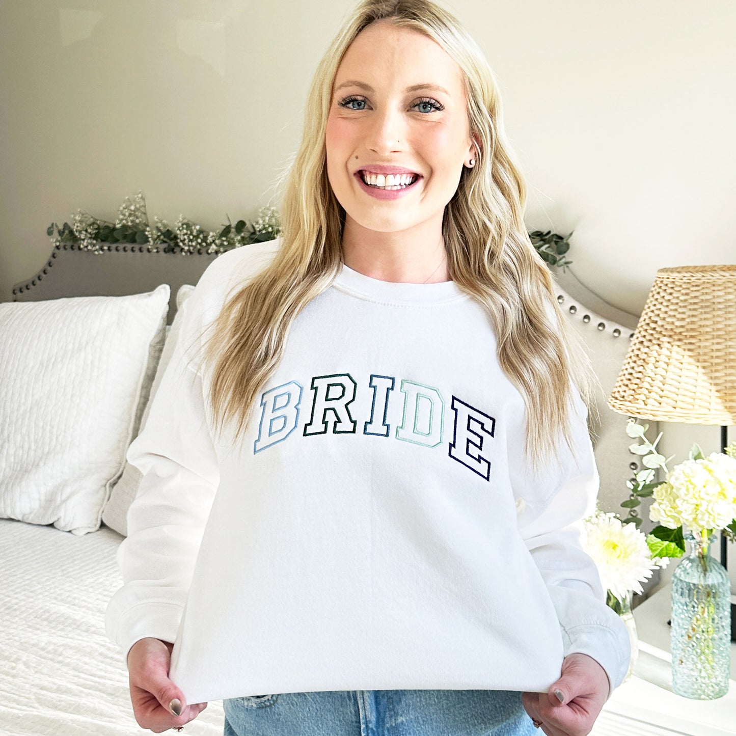 young woman wearing a white pullover crewneck sweatshirt with bride embroidered in an athletic block outlined font in green and blue threads.
