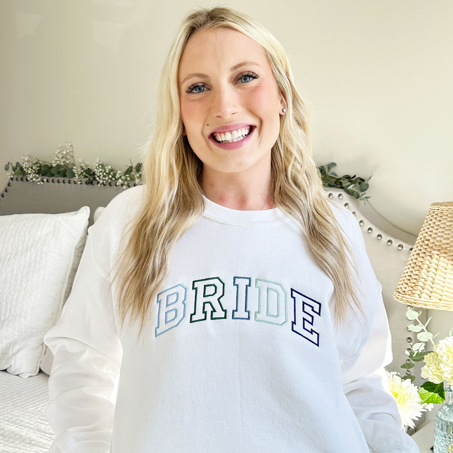 bride wearing a white crewneck sweatshirt with bride embroidered across the chest in a varsity font and blue and green thread