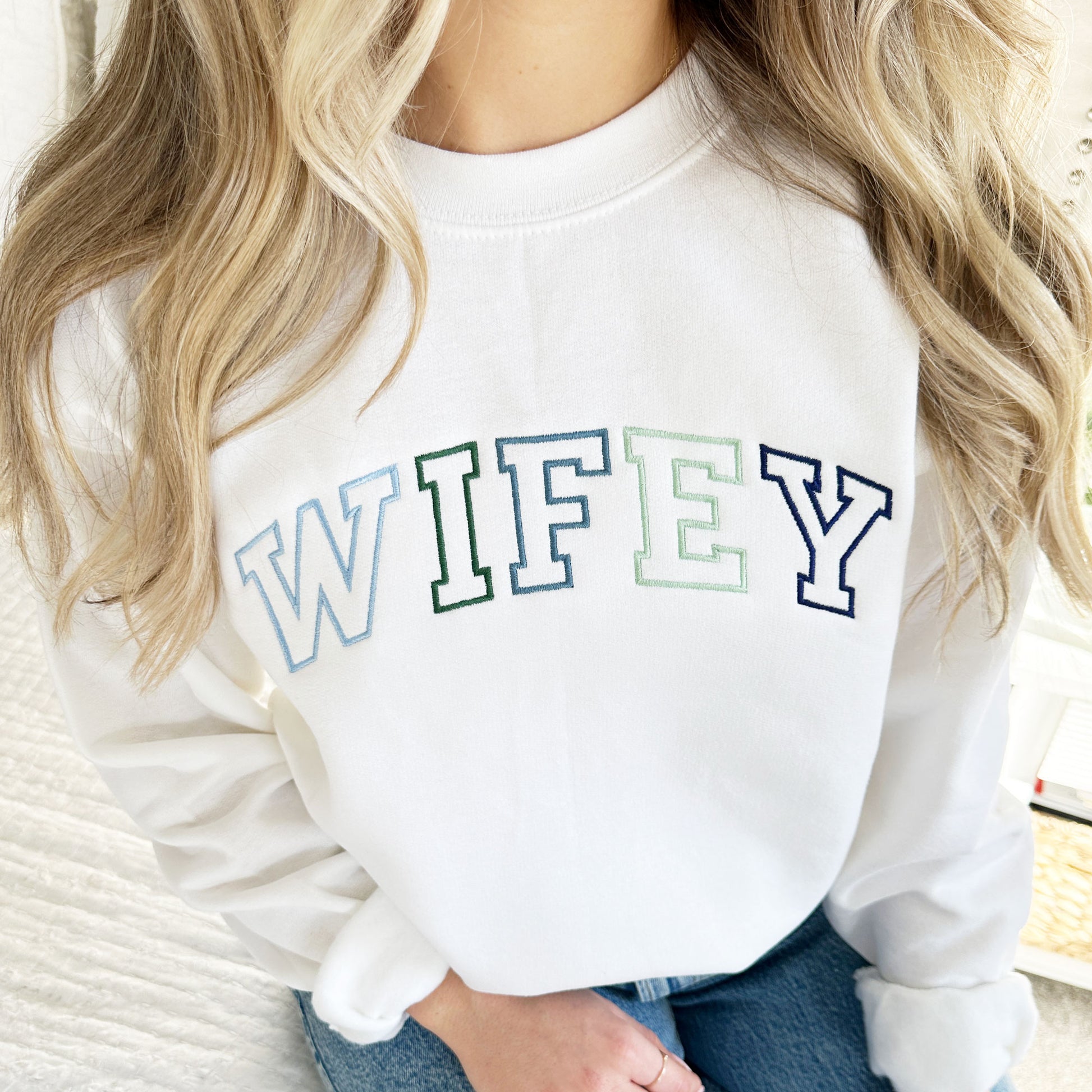 woman wearing a white sweatshirt with a blue green embroidered wifey design