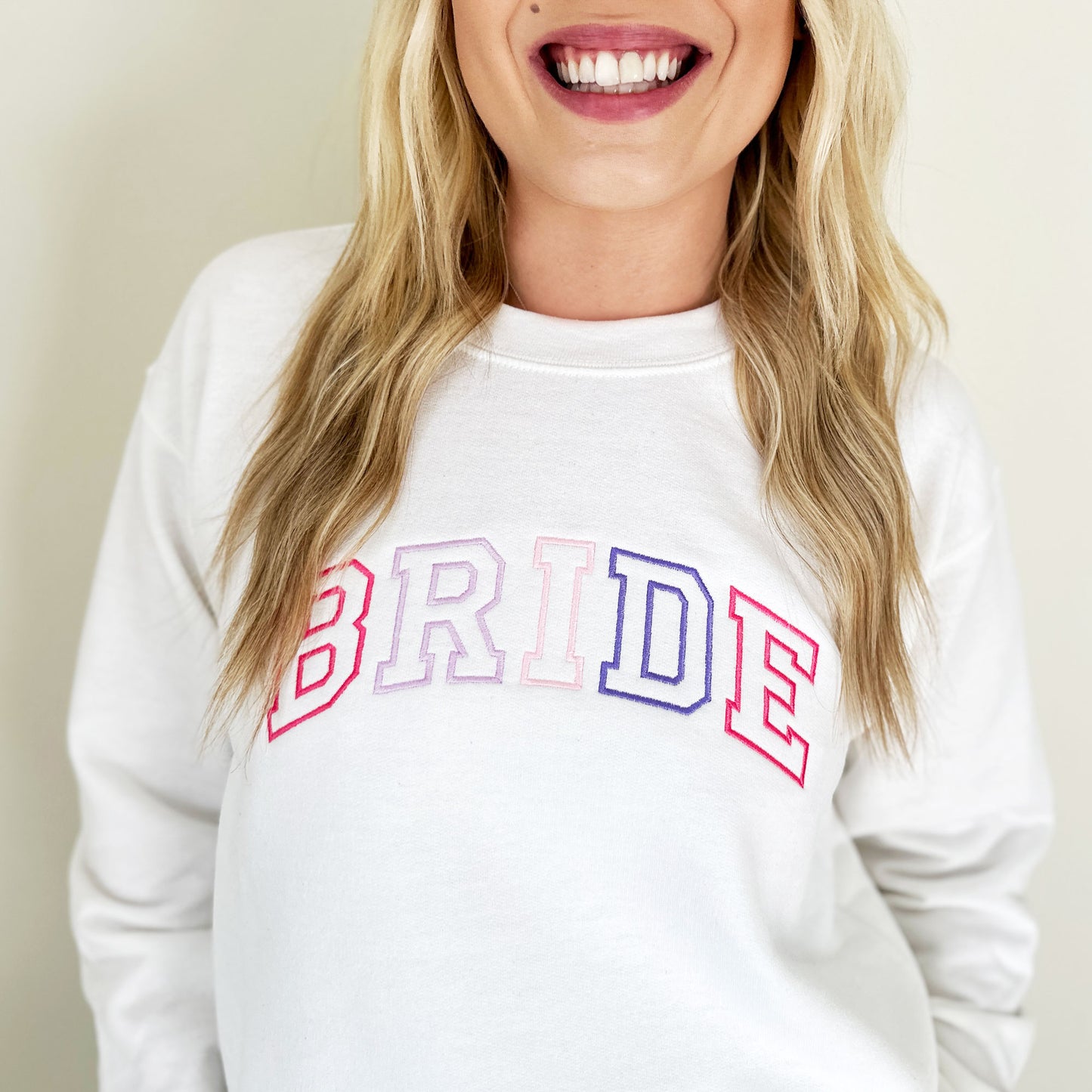 woman wearing a white sweatshirt with a custom BRIDE embroidered design across the chest in a cute pink and purple thread pattern