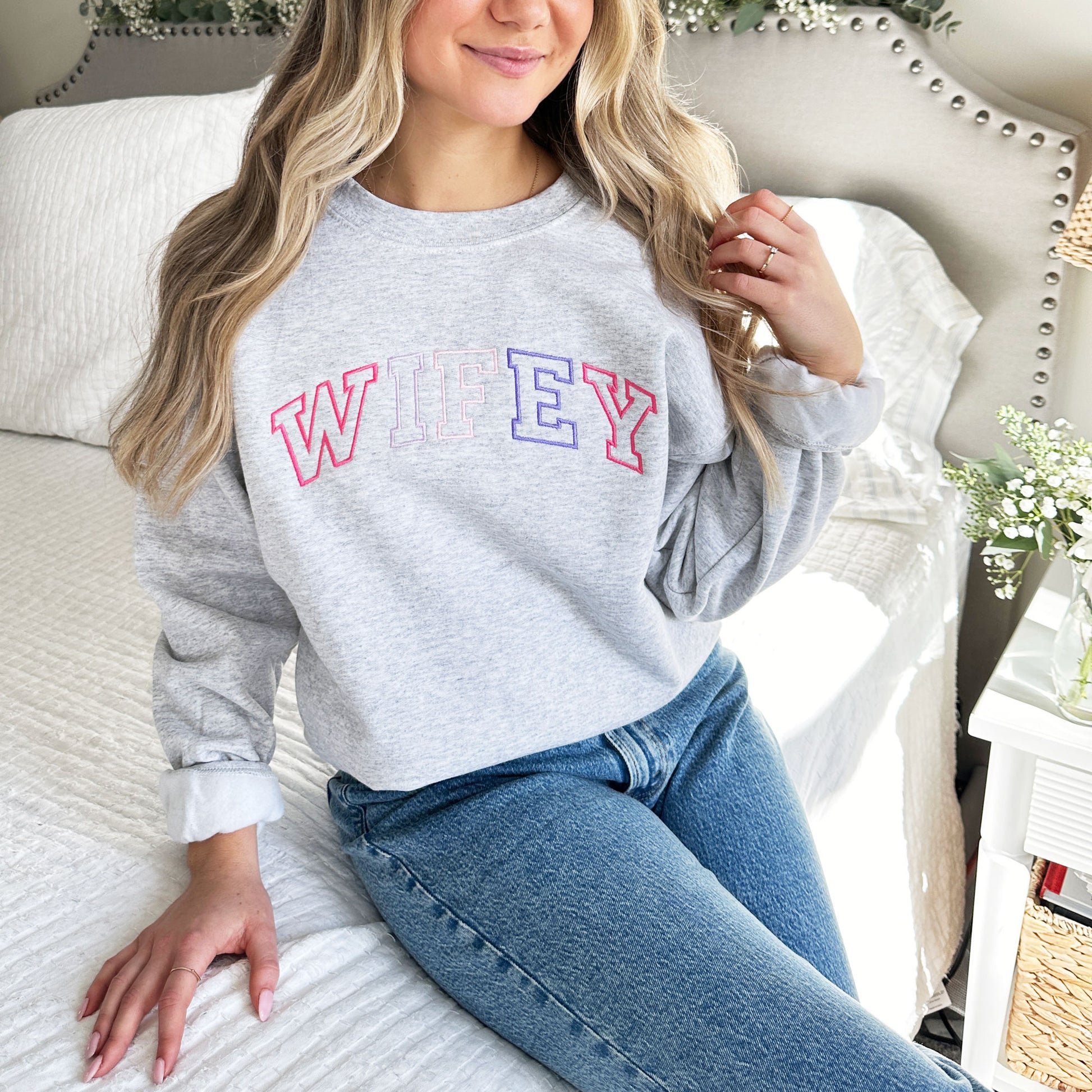 woman sitting on the edge of the bed getting ready for her wedding wearing an ash gray sweatshirt with wifey embroidered in a large athletic block font across the chest