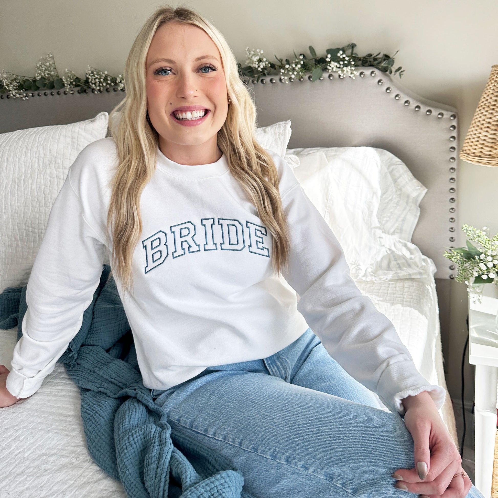 woman wearing a white pullover crewneck sweatshirt with bride embroidered in a blue thread across the font center chest