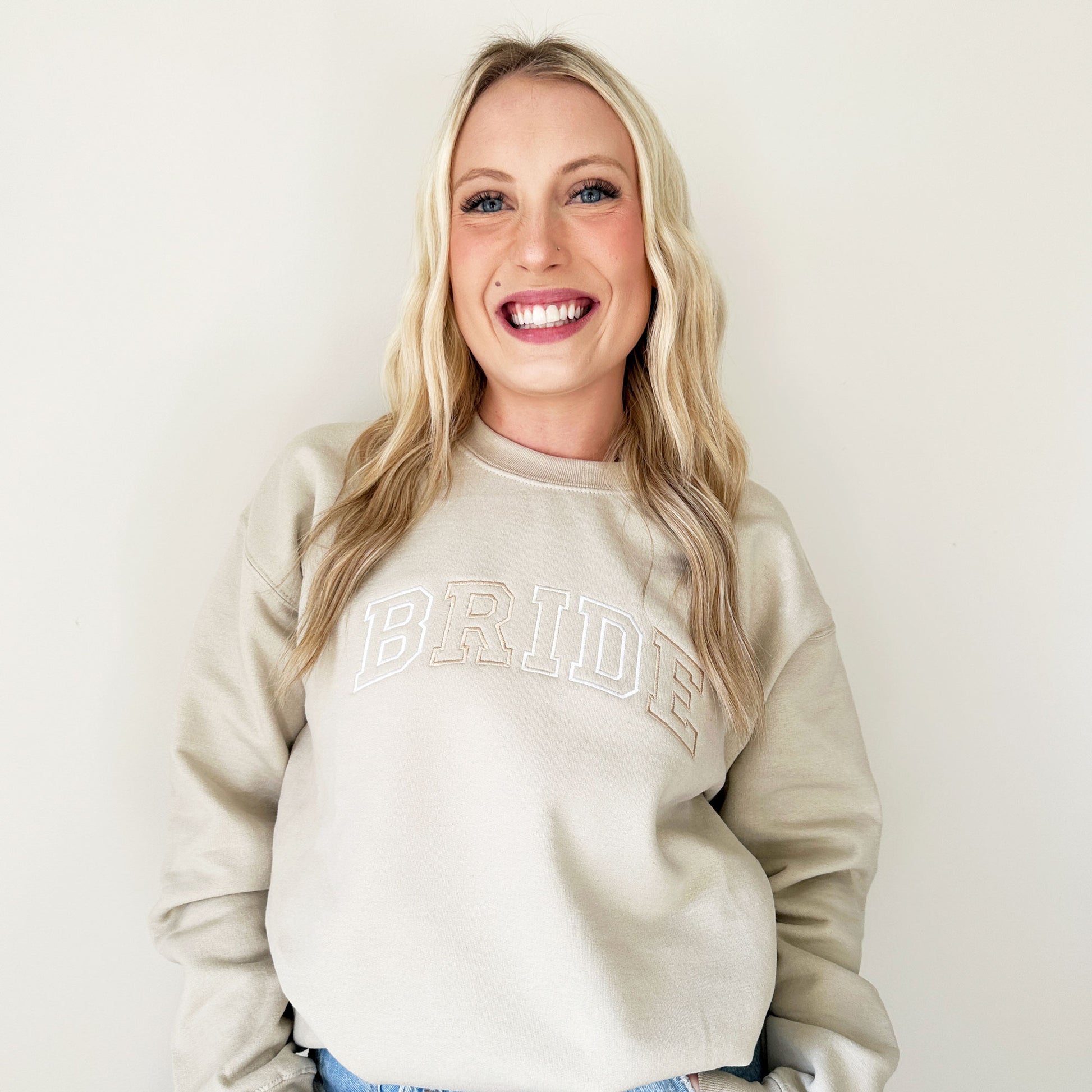 young woman wearing a light beige pullover sweatshirt with BRIDE embroidered across the chest in a neutral thread combination
