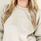 close up of a BRIDE design embroidered in an atheltic block open font across the chest of a sand sweatshirt in various neutral thread colors