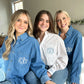 3 young women sitting n a bed modeling denim button up shirts with monogram embroidery on the pocket in white thread