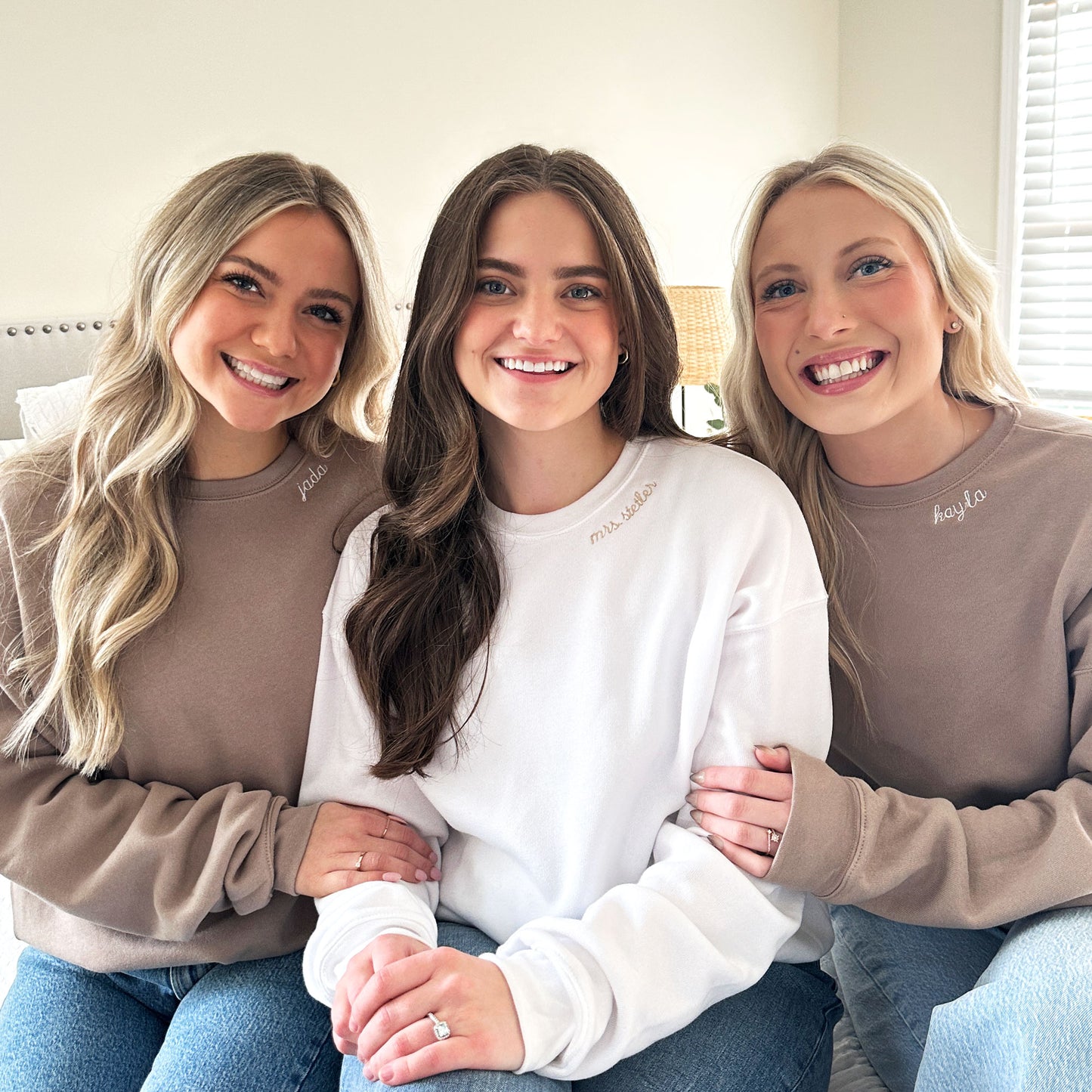 3 young women wearing crewneck sweatshirts for a bridal photo. The bride is wearing a white crewneck sweatshirt with custom name embroidered in camel thread on the neckline. the bridesmaids are wearing a tan crewneck sweatshirt with names embroidered in white on the neckline 