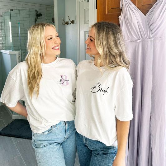 a bride and her bridesmaid wearing oversized white t-shirts with custom left chest printed designs. the bride's shirt has bride in a script font on the left chest and the bridesmaid has an initial and name  print on the left chest.
